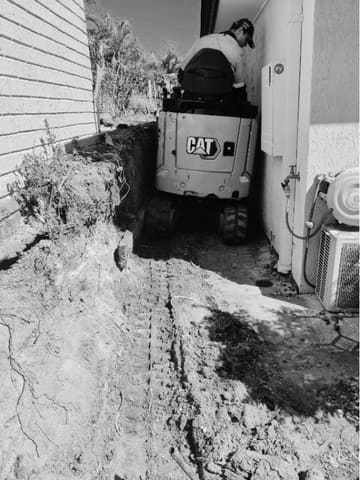 Mini excavator digging a trench down a narrow side path between a house and the boundary in Forest Lake